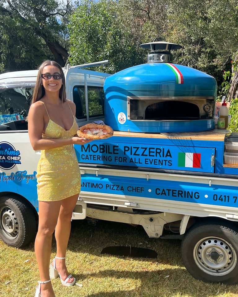 Party guest holding a wood-fired pizza beside the blue pizza truck