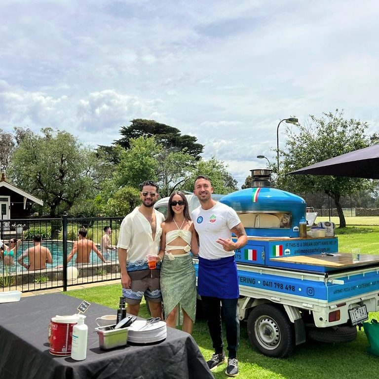Hosts relaxing with guests beside the blue pizza truck at a garden party